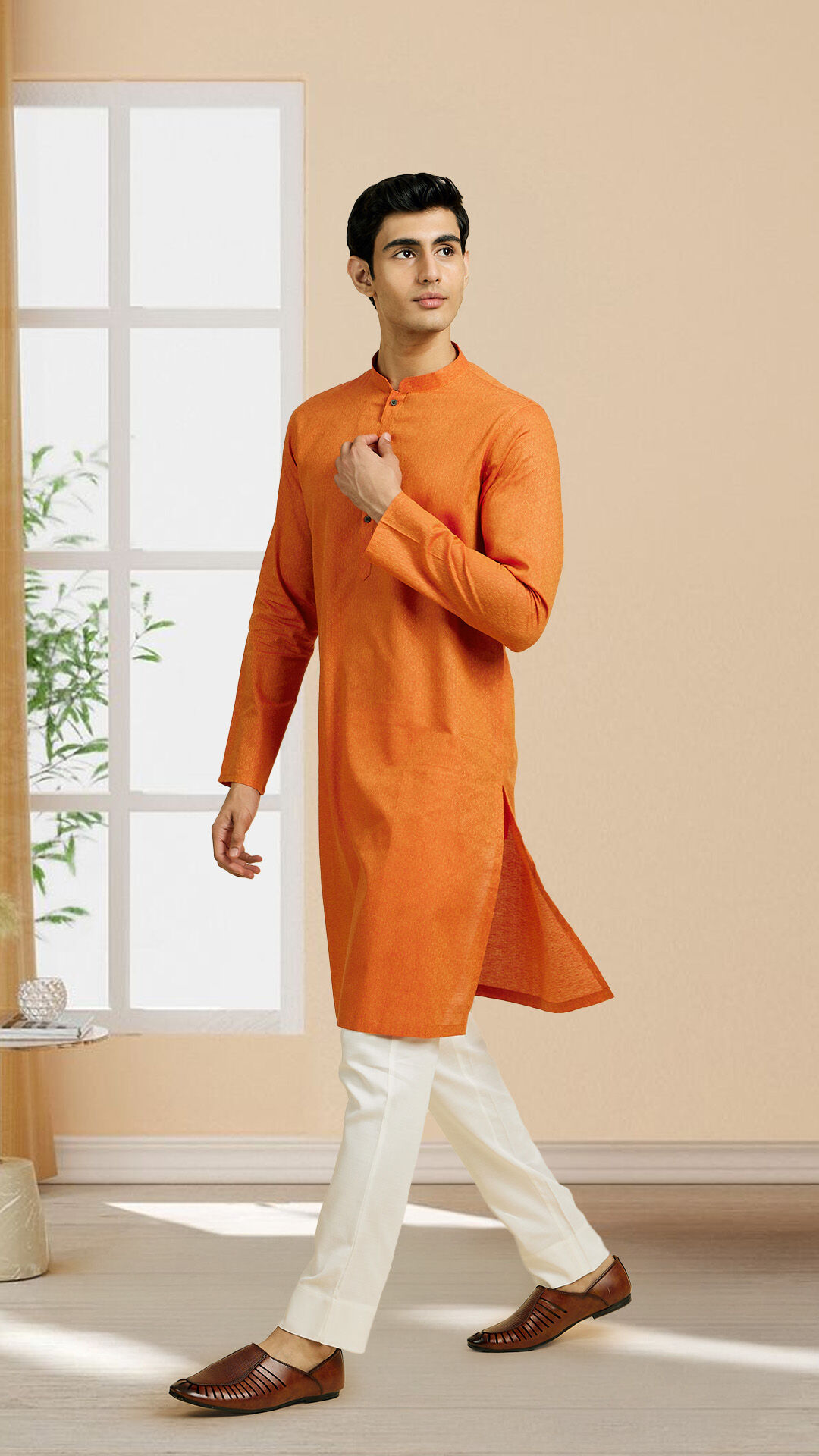 Man wearing an orange cotton kurta with white trousers, walking indoors with natural light streaming through a window, highlighting breathable and stylish summer ethnic wear.